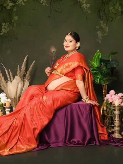 A graceful solo portrait of a mom-to-be in a beautiful red silk saree. Holding a peacock feather, she sits elegantly in our studio setup designed for traditional photoshoots.