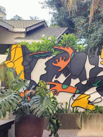 This colorful outdoor wall installation is made of layered, painted metal. The abstract, biomorphic shapes are designed to interact with the surrounding foliage, bringing a sense of playful energy and 'fun and frolic' to the exterior space.