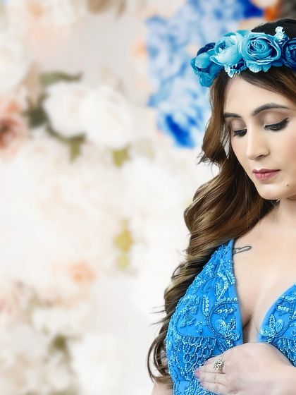 A close-up detail shot from a maternity session. The focus is on the mother's serene expression, her blue floral crown, and the intricate beading of her gown.