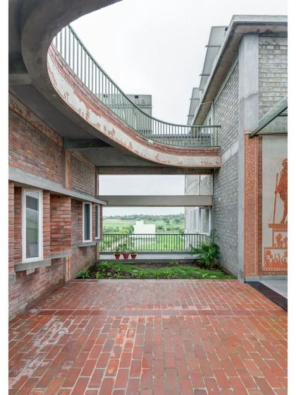 A curved bridge connects two parts of the ASA office, framing a view of the landscape. The building uses a mix of exposed fly ash blocks and warmer brick tones, with murals adding a touch of local art.