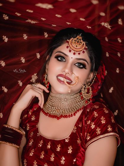 A glamorous bridal portrait where the bride is looking directly at the camera. The rich red background complements her velvet lehenga and traditional nath, creating a bold and beautiful look.
