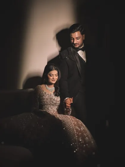 A quiet, tender moment captured with artistic lighting. The groom holds his bride's hand as she sits, their connection highlighted by a single beam of light in a darkened room.