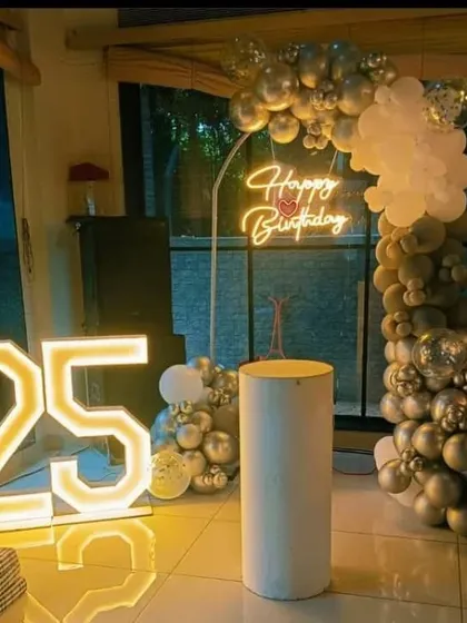 A 25th birthday party with a silver and white balloon arch, a 'Happy Birthday' neon sign, and a large '25' marquee light.