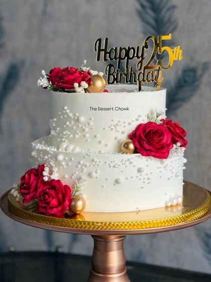 A beautiful two-tier cake celebrating a 25th birthday. The design features classic white buttercream, delicate sugar pearls, and is adorned with deep red roses for a touch of elegance.