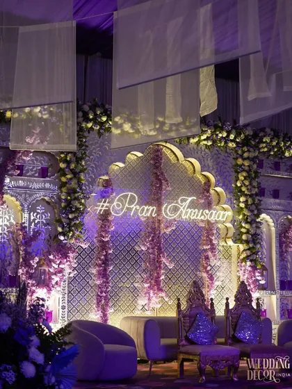 The couple's neon hashtag glowing against the lavender-lit jaali backdrop. Cascading purple flowers add a touch of softness and romance to this modern and personalized stage design.