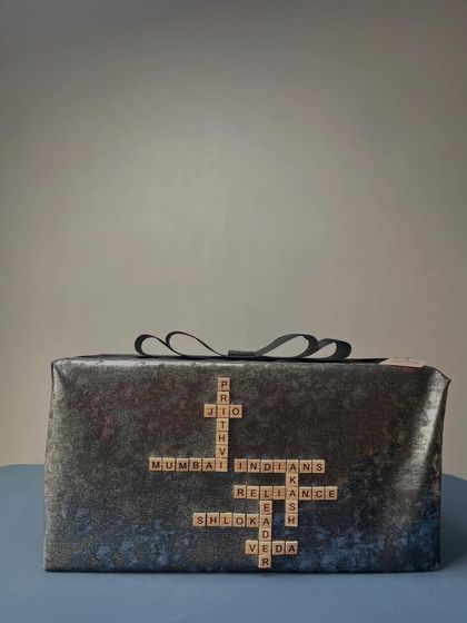 A truly personal touch, this gift is wrapped in dark, textured paper and features a custom Scrabble design with the names of family members and important connections, like 'Prithvi', 'Shloka', and 'Mumbai Indians'.