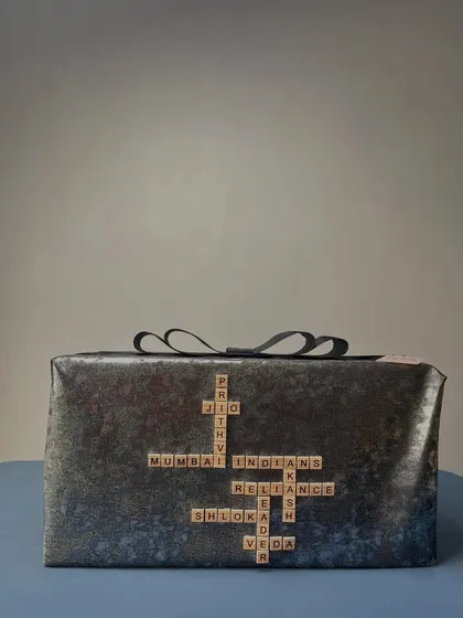 A truly personal touch, this gift is wrapped in dark, textured paper and features a custom Scrabble design with the names of family members and important connections, like 'Prithvi', 'Shloka', and 'Mumbai Indians'.