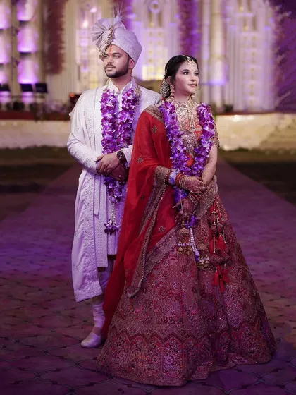 A formal portrait of the bride and groom on their wedding day, adorned with beautiful purple garlands.
