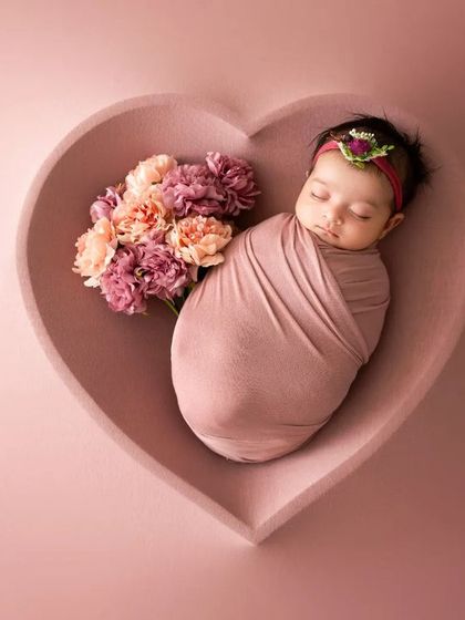 A newborn swaddled in pink sleeps in a heart-shaped bowl, accented with a bouquet of flowers. This setup is perfect for a classic and beautiful baby portrait.