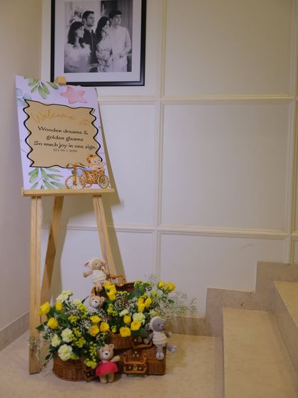 A welcoming corner at the baby shower, featuring a custom sign on an easel, wicker baskets filled with flowers and crochet toys, and a framed photo of the happy couple.