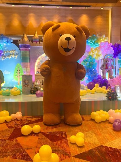 Our brown Teddy Bear mascot stands ready to party in a room filled with colorful balloons, perfectly matching the festive birthday atmosphere.