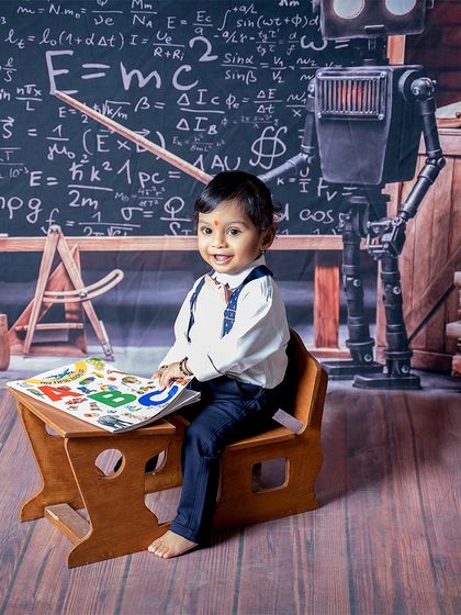 A little scholar with a bright future. This happy baby is enjoying his book at a tiny desk, with a robot and chalkboard adding to the fun, educational theme.