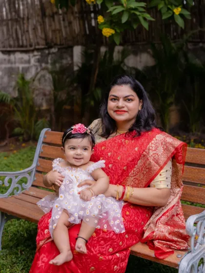 A mother and her smiling daughter share a sweet moment on a park bench. The vibrant red of her saree looks stunning against the garden backdrop.
