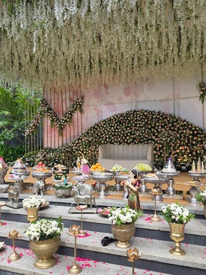 A grand engagement stage with a massive floral arch made of dark roses and a ceiling of hanging white flowers. The multi-level stage is filled with brass and silver offering vessels.