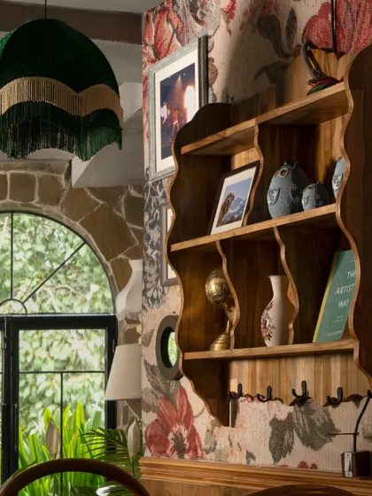 A scalloped wooden shelf holds a collection of curiosities, set against a backdrop of floral and stone textures. The fringed green lampshade adds a touch of playful maximalism to this eclectic corner.