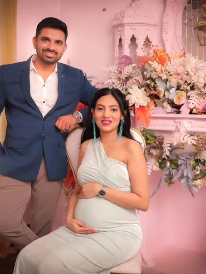 A stylish couple pose during their maternity session. The setup includes an elegant chair for the mom-to-be, placed beside the beautifully decorated pink fireplace prop, offering a variety of posing options.