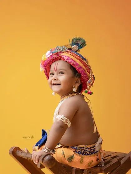A bright and cheerful portrait of a baby as Krishna, looking over his shoulder with a sweet smile. The solid yellow background highlights the colorful turban and joyful expression.
