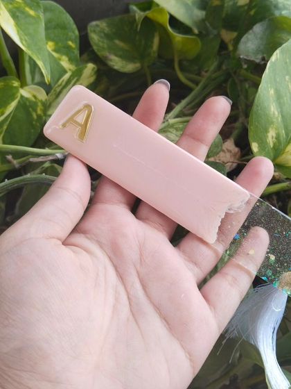 Holding the pastel pink bookmark with the 'A' initial. This view shows the two-tone design with a glittery clear section at the bottom.