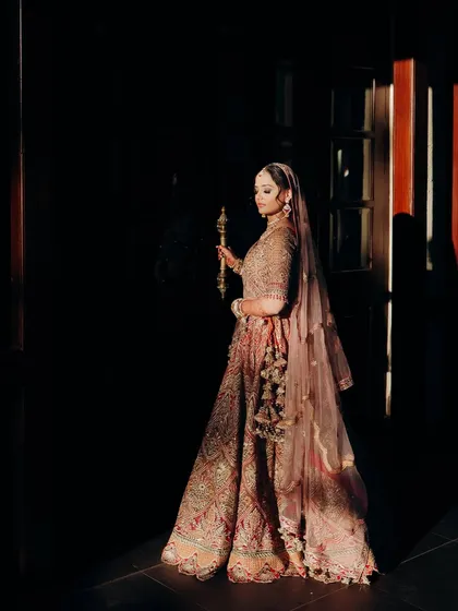 A dramatic and artistic shot of the bride. The lighting highlights the silhouette of her lehenga and the regal elegance of her makeup and posture.