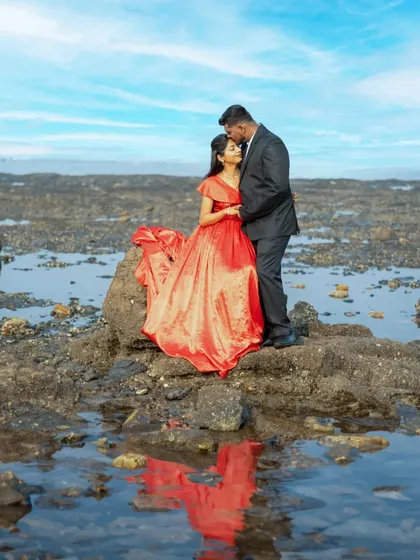 A beautiful pre-wedding portrait with a dramatic sky and water reflection. The bride's vibrant red gown contrasts beautifully with the serene landscape.