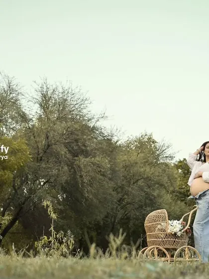 A modern and casual outdoor shot. The mom-to-be shows off her bump in jeans and a white shirt, standing next to a rustic baby pram in a wide-open field.