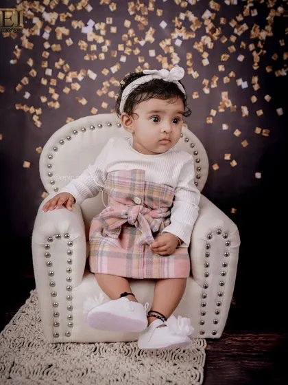 This little girl looks thoughtful and sweet in her plaid dress, seated in a mini armchair against a gold confetti backdrop for a touch of sparkle.
