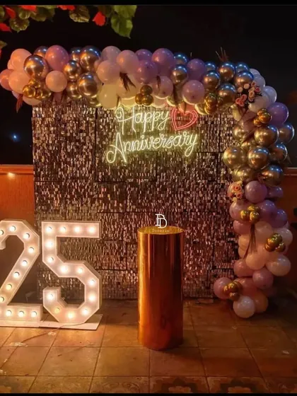 A stunning 25th-anniversary celebration on a terrace. The decor features a silver sequin wall with a 'Happy Anniversary' neon sign, large '25' marquee numbers, and a balloon arch in pastel and metallic shades.