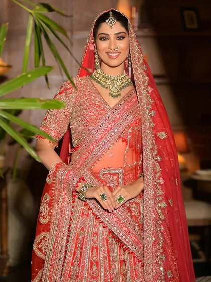 A smiling bride in a red lehenga, her look perfected with a green Kundan choker, maang tikka, and nath. A truly classic and elegant bridal style.