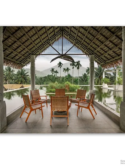 Looking out from a tranquil seating area at Maayaa Resorts in Anaikatti, this photograph frames the majestic mountains through stone pillars. The thatched roof and serene infinity pool create a seamless connection between the architecture and the breathtaking natural landscape, promising a peaceful escape.