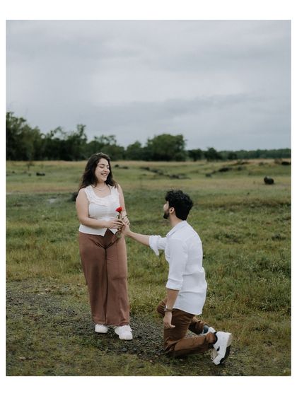 A beautiful, full-length shot of a proposal moment in a green field. The composition gives a sense of place and captures the gentle, romantic gesture in a natural setting.