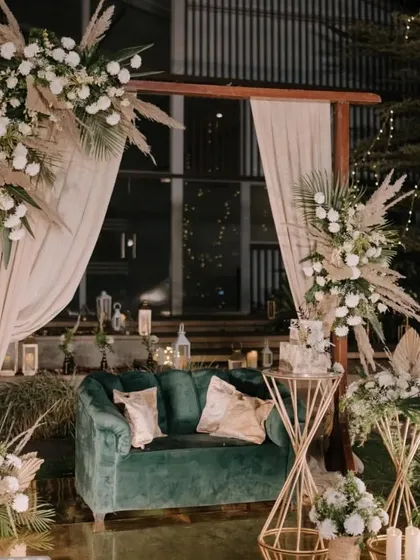 For a modern and chic reception, we designed this sophisticated lounge area. The setup features a plush emerald green sofa, complemented by dried pampas grass and white floral arrangements on geometric gold stands, creating a look of understated luxury.