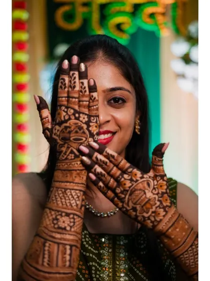 The bride playfully peeks through her hands, showing off her elaborate henna design. This is a fun and classic pose for a mehendi ceremony.