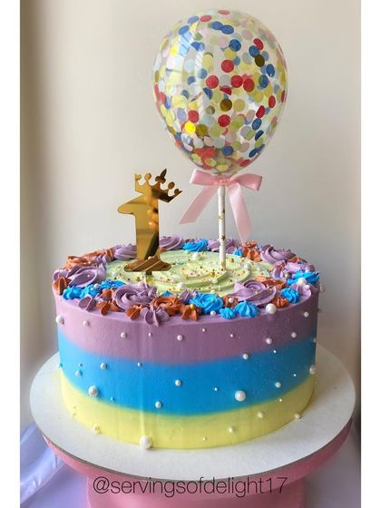 A colorful first birthday cake featuring a confetti balloon topper. The rainbow buttercream stripes make it a cheerful and festive choice for a celebration.