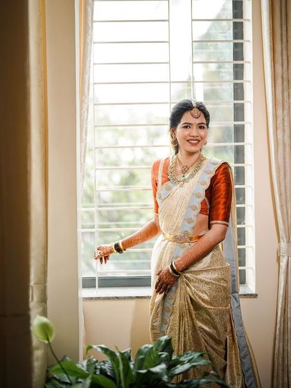 A beautiful, naturally lit portrait of a bride in a classic off-white and rust-orange silk saree. The makeup is kept soft and natural to enhance her features, creating a look of pure elegance.