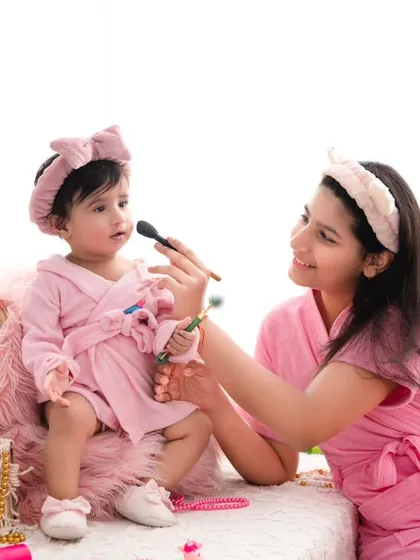 A playful spa day for mother and daughter. Dressed in matching pink robes, they share a fun moment with makeup brushes, creating a unique and adorable memory.