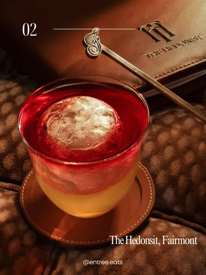 A beautifully layered cocktail from The Hedonist. The lighting perfectly captures the separation of colors and the large, clear ice sphere. The branded menu in the background adds context and reinforces the bar's identity.