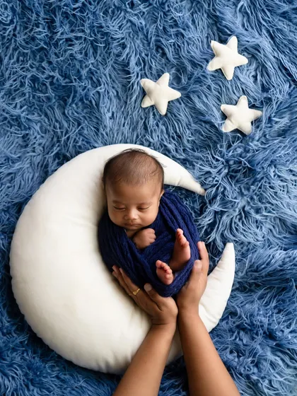A newborn wrapped in deep blue, held in a crescent moon prop against a furry blue backdrop with soft white stars. This shot is a beautiful example of a celestial-themed photoshoot.