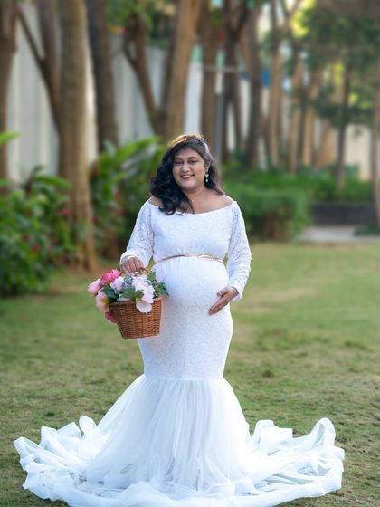A lovely outdoor shot of a mom-to-be in a white lace gown, holding a basket of flowers.