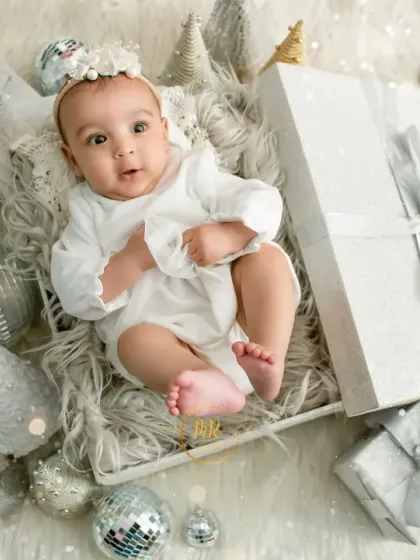 A baby girl in a white outfit surrounded by silver and white Christmas presents and ornaments for a magical holiday photo.