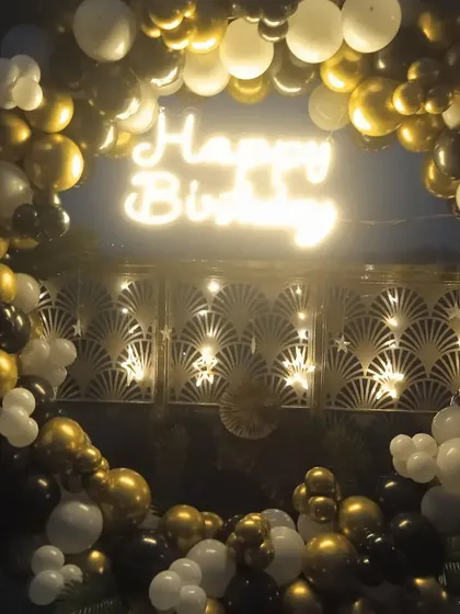 An outdoor evening party setup with a black, gold, and white balloon ring. The 'Happy Birthday' neon sign glows beautifully against the decorative screen backdrop.