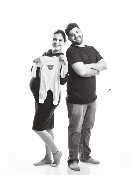 A fun and casual black and white portrait. The couple stands back-to-back, with the mom-to-be proudly holding up a baby onesie, capturing their happy and relaxed dynamic.