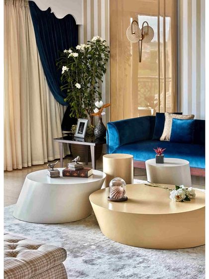 A dramatic and elegant living room composition. The asymmetrical center table cluster, deep blue velvet sofa, and a gorgeous custom pelmet framing the curtains create a perfect balance of style and sophistication.