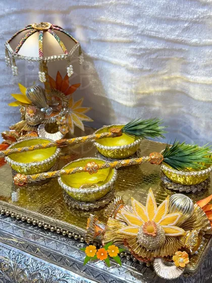 A close-up of a custom haldi platter. The yellow bowls are decorated with gota work and sit on a silver tray, accompanied by a small decorative umbrella, ready for the ceremony.