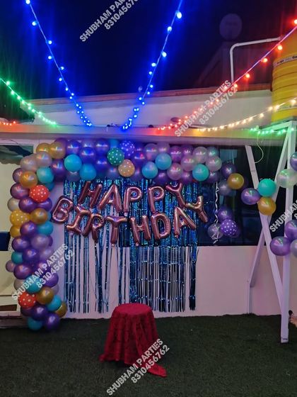 A rooftop birthday party at night with colorful string lights and a balloon backdrop.