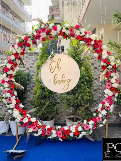 An elegant and rustic "Oh Baby" backdrop for a baby shower. A large ring made of fresh pink and white flowers creates a beautiful frame for photos, perfect for an outdoor or garden setting.