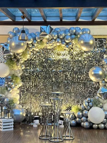 A sophisticated birthday setup with a silver sequin wall. The decor is accented with a full arch of silver and white balloons, star-shaped foil balloons, and modern wire-frame pedestals.