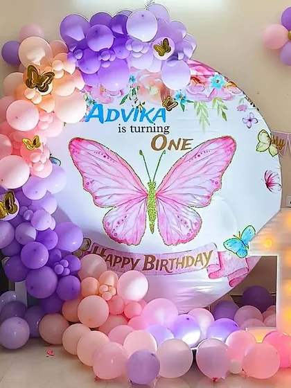 A vibrant butterfly theme first birthday setup perfect for a photo corner. It includes a large circular backdrop with a central butterfly graphic, a multi-colored balloon arch in pink and purple, and a light-up number '1' prop.