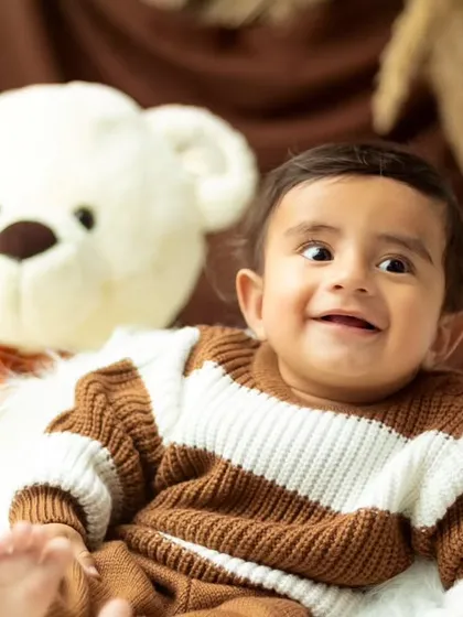 A cute and quirky expression from a baby boy in a cozy sweater, sitting next to his teddy bear friend.