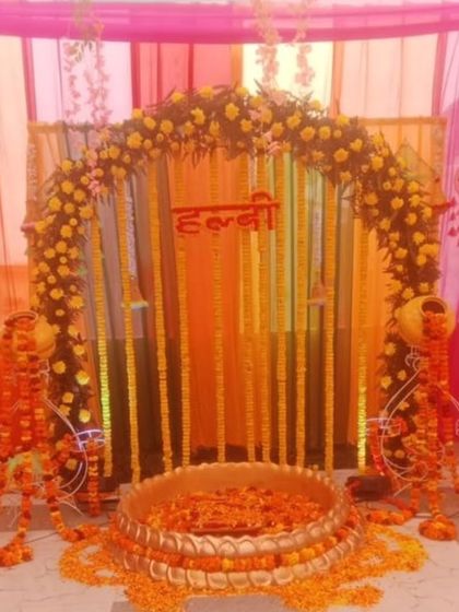 A traditional Haldi ceremony setup with a floral arch and a large brass urli. The decor uses marigold garlands and decorative matkas on stands, perfect for a home-based function.