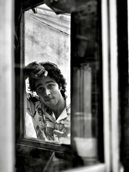 A black and white portrait of Zahan Kapoor seen through a window. The reflection and framing add a layer of complexity and emotion to the shot.